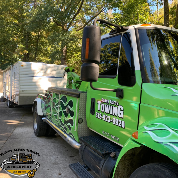 A green tow truck from Sunny Acres Towing with a white travel trailer in Greenville, IN.