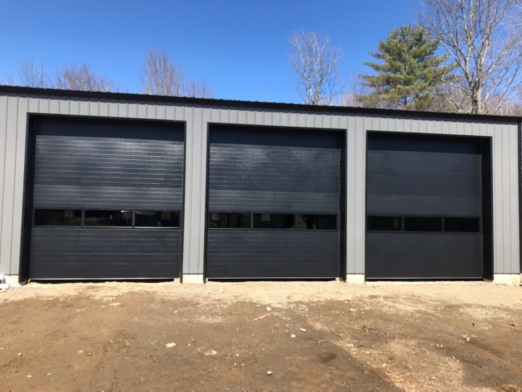 Three black industrial garage doors with windows installed on a grey metal building by Ski Door, Inc. in Center Rutland, VT.