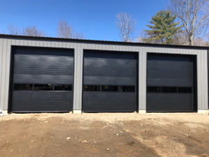 Three black industrial garage doors with windows installed on a grey metal building by Ski Door, Inc. in Center Rutland, VT.