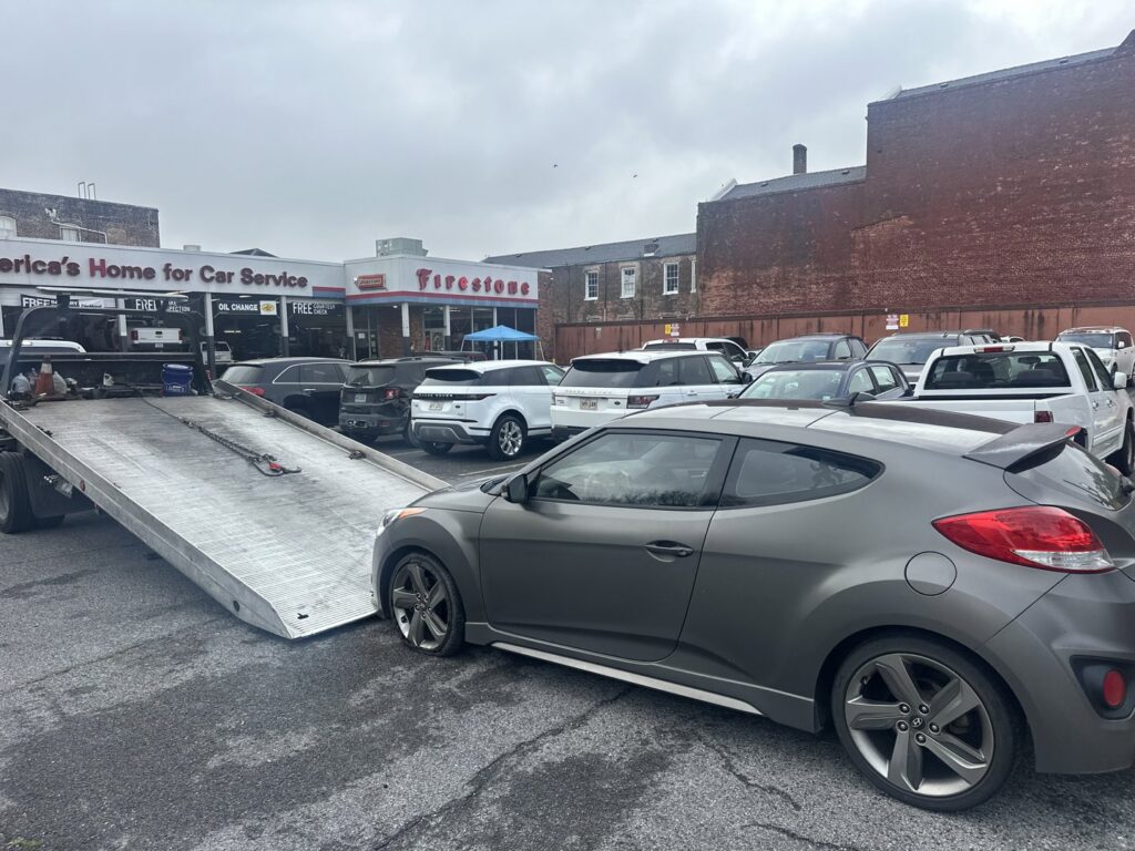 A grey Hyundai Veloster parked next to a TJ Towing LLC flatbed truck, ready for towing in New Orleans, LA.