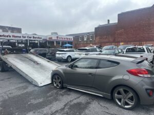 A grey Hyundai Veloster parked next to a TJ Towing LLC flatbed truck, ready for towing in New Orleans, LA.