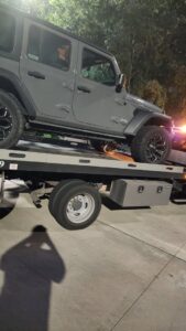 A grey Jeep Wrangler being towed on a flatbed tow truck by Jay Towing Services at night in Houston, TX.