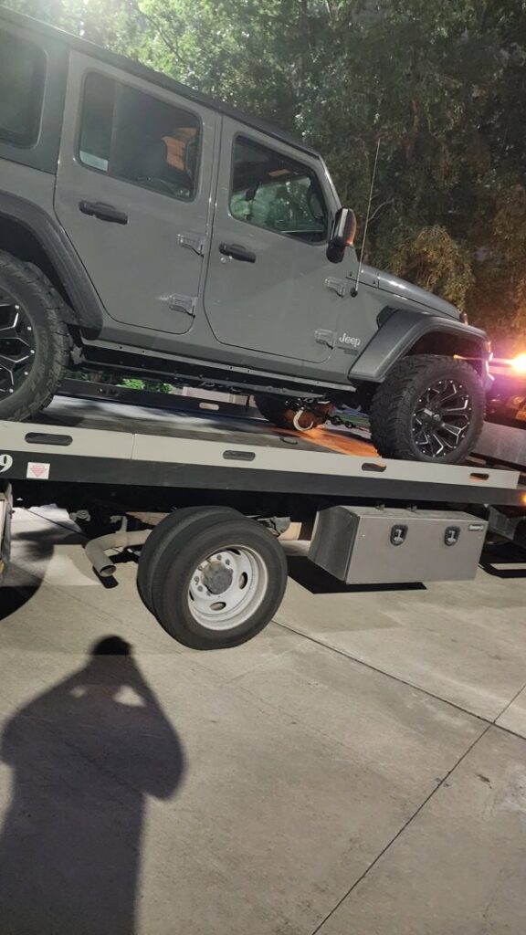 A grey Jeep Wrangler being towed on a flatbed tow truck by Jay Towing Services at night in Houston, TX.