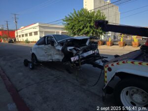 A heavily damaged white car being towed by a tow truck from Black Rhino Towing in Denver, CO.