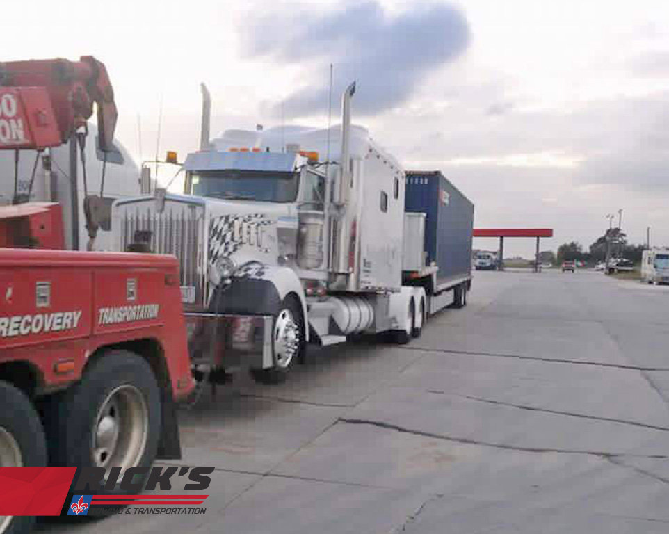 A heavy-duty tow truck from Rick's Towing and Recovery in Lafayette, LA, assisting a semi-truck with a blue container trailer.