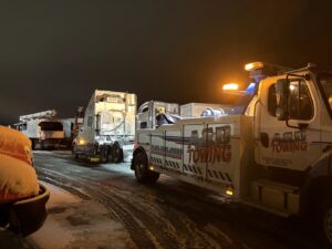 A heavy-duty tow truck from A and D Towing is recovering multiple semi-trucks on a snowy night in Little Ferry, NJ.