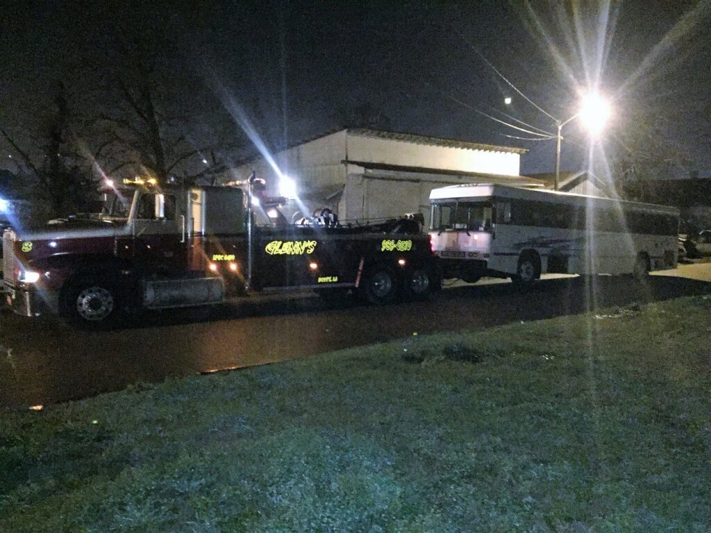 A heavy-duty tow truck from Glenn's Towing & Recovery Inc towing a large white bus at night in Lafayette, LA.