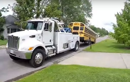 A heavy-duty tow truck from Primo Towing towing a yellow school bus down a street in Anderson, IN.