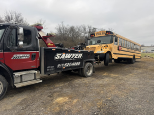 A heavy-duty tow truck from Sawyer Towing is actively towing a yellow school bus in Cincinnati, OH.