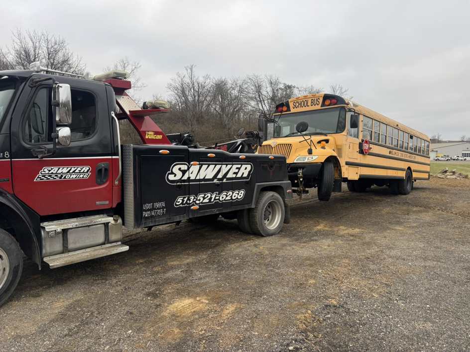 A heavy-duty tow truck from Sawyer Towing is actively towing a yellow school bus in Cincinnati, OH.
