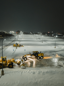 An aerial view of multiple heavy equipment machines clearing a large snowy parking lot at night for Elcor Construction in Rochester, MN.