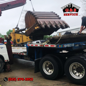 G & S Towing Service, Inc. crew lifting a heavy excavator bucket onto a flatbed trailer in Des Moines, IA.