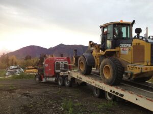 Heavy equipment transport showing a loader being driven onto a flatbed trailer by AK Car Crushing & Recycling & ACCR Towing in Wasilla, AK.