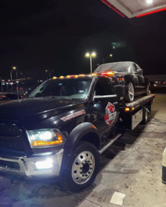 A Heavy Hookz Towing flatbed truck with a car loaded, parked at a gas station at night in Fort Worth, TX.