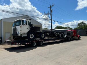 A red Elite Towing of Minnesota semi-truck transporting a white truck chassis on a flatbed trailer in St Paul, MN.