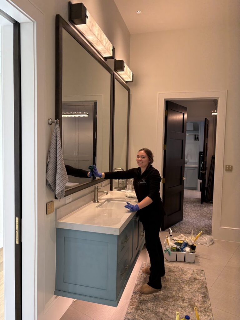 A Kleen Slate house cleaner smiling while cleaning a large bathroom mirror and vanity with a cloth in Shakopee, MN.