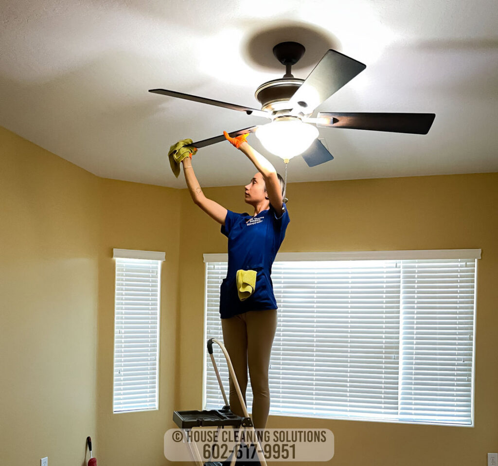 A house cleaner from House Cleaning Solutions in Mesa, AZ, is cleaning a ceiling fan from a step ladder.