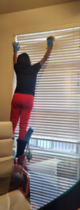 A house cleaner wearing gloves, standing on a step stool, cleaning window blinds for Creativity Cleaning LLC in Detroit, MI.