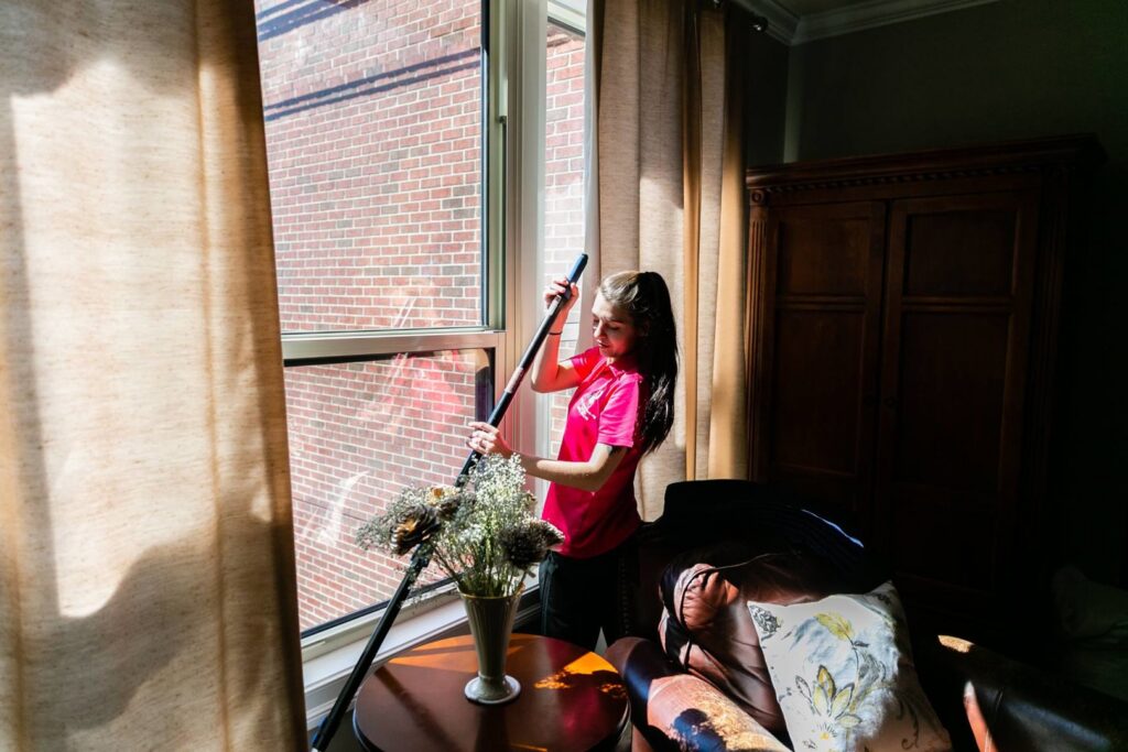 A house cleaner in a pink shirt using a long-handled tool to clean a window at a client's home by New Image Cleaning Service LLC in Richmond, VA.