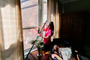 A house cleaner in a pink shirt using a long-handled tool to clean a window at a client's home by New Image Cleaning Service LLC in Richmond, VA.