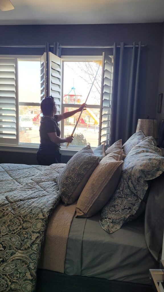 A house cleaner from ECO Friends cleaning service dusting a bedroom window with a long tool in Carson City, NV.