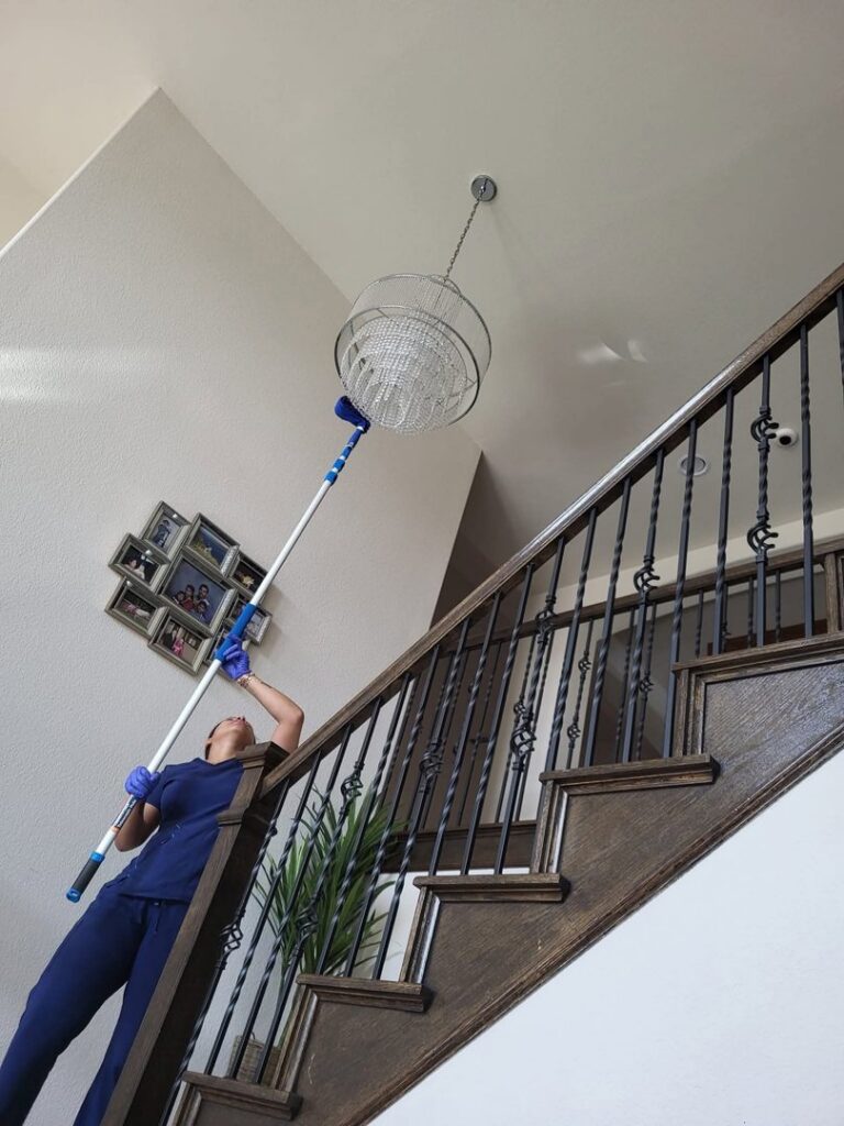 A house cleaner using an extension pole to dust a high chandelier, demonstrating professional cleaning by Supreme Cleaning Services in Farmers Branch, TX.