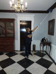 A house cleaner wearing gloves, dusting high corners in a room with a chandelier for Creativity Cleaning LLC in Detroit, MI.