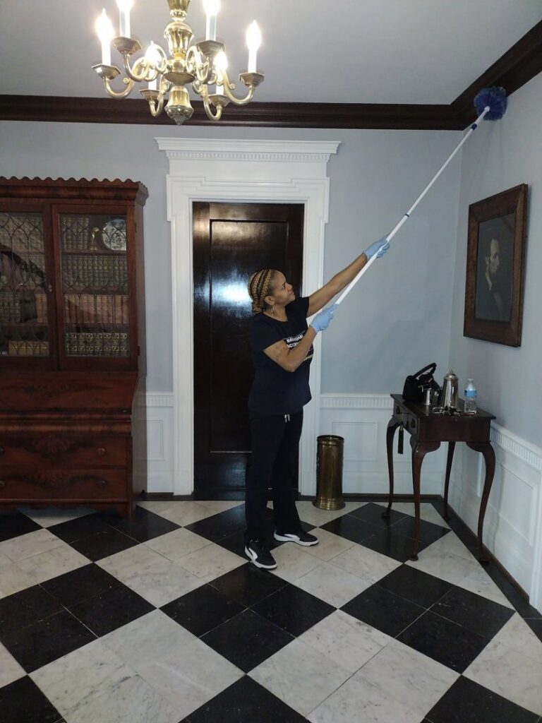 A house cleaner wearing gloves, dusting high corners in a room with a chandelier for Creativity Cleaning LLC in Detroit, MI.