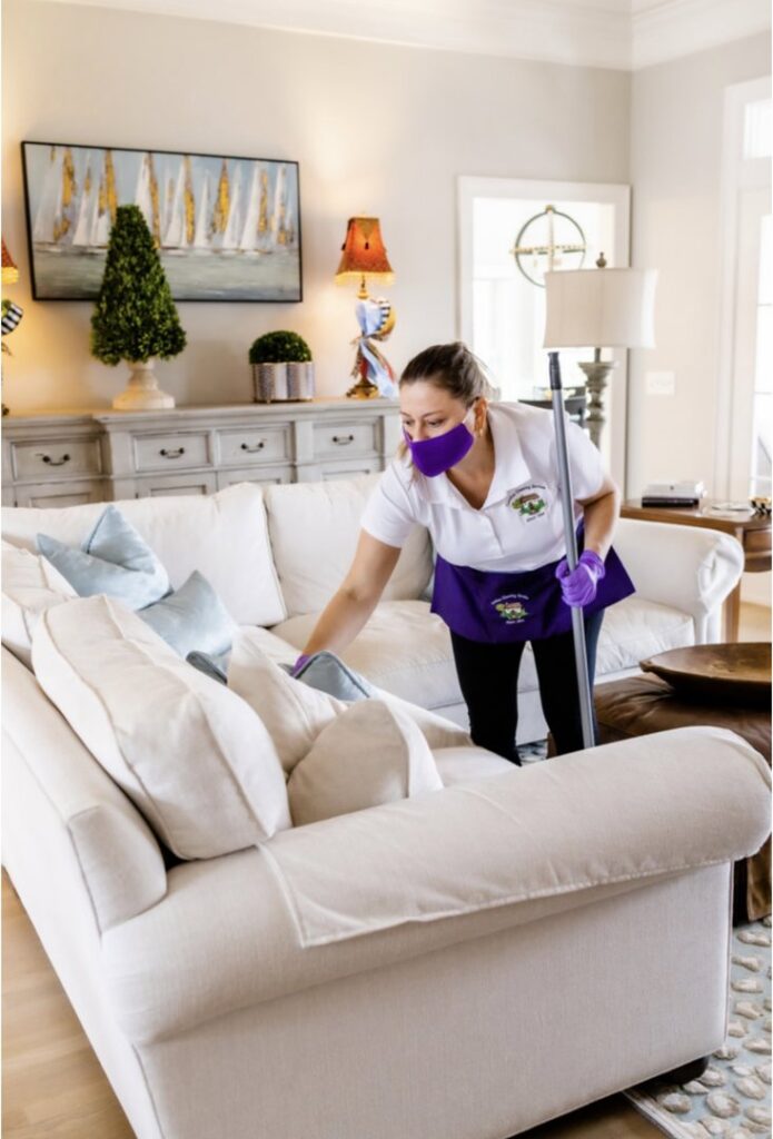 A house cleaner from Carolina Cleaning Services in Rocky Mount, NC, dusting a white sofa in a bright living room.