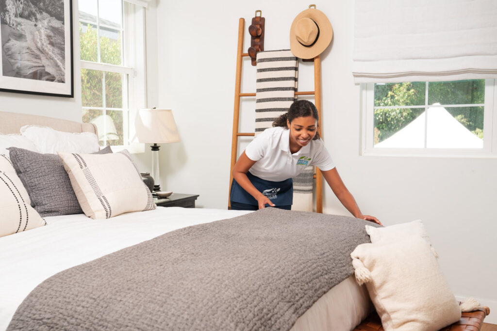 A house cleaner from The Cleaning Authority - Bowie, MD, neatly making a bed in a bright bedroom.
