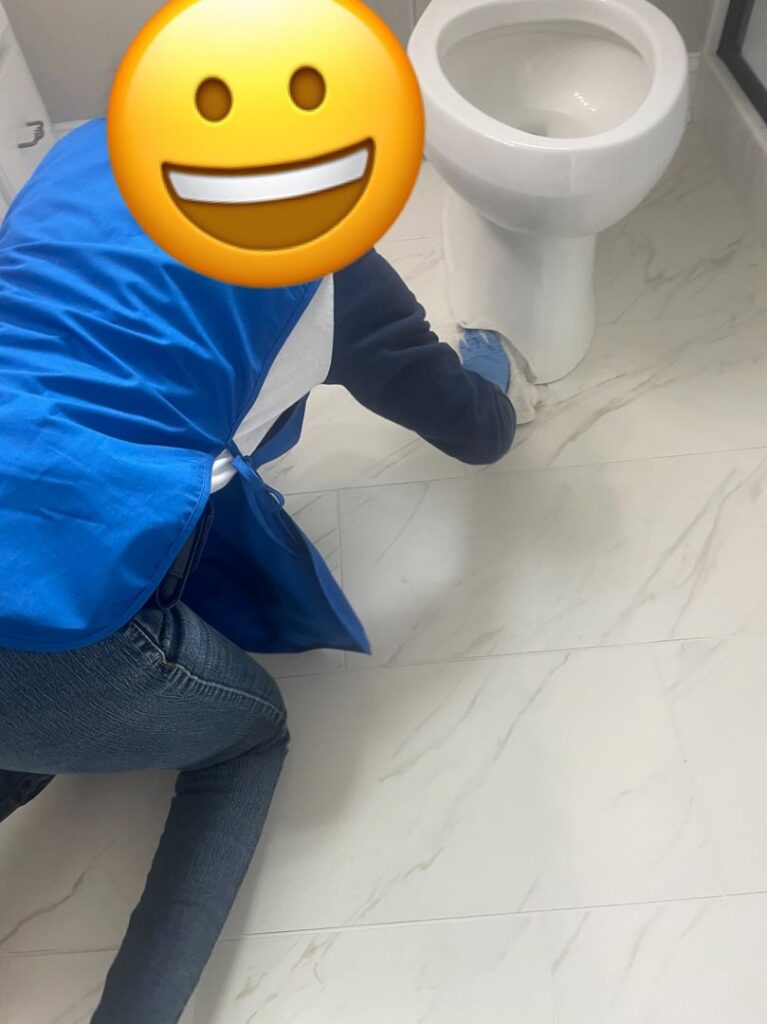 A house cleaner in a blue apron meticulously scrubbing around a toilet in a bathroom for Skedaddle Cleaning Services in Eufaula, AL.
