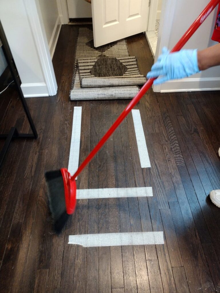 A house cleaner wearing gloves, sweeping a wooden floor with a broom for Creativity Cleaning LLC in Detroit, MI.