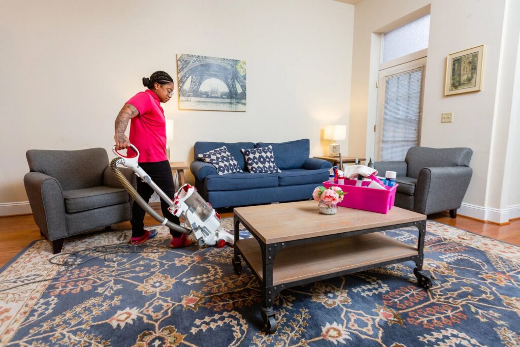 A house cleaner vacuuming a large patterned rug in a living room, with cleaning supplies on the coffee table, by New Image Cleaning Service LLC in Richmond, VA.