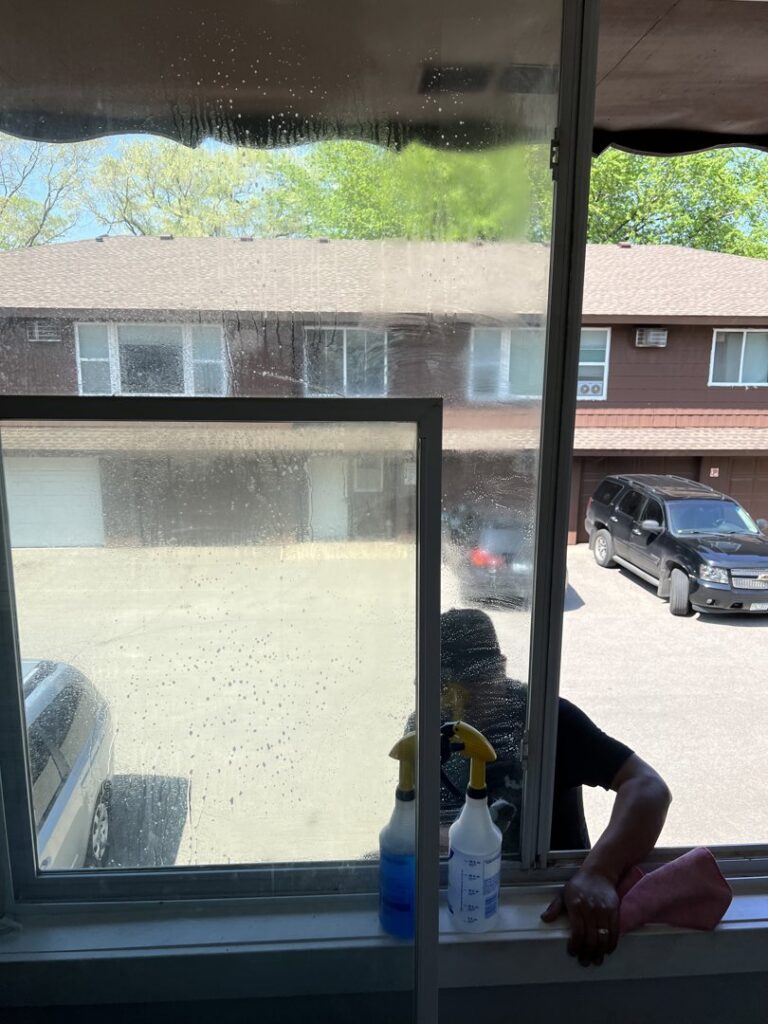 A house cleaner actively washing a window with spray bottles and a cloth, demonstrating window cleaning services by Little Cubs Cleaning Services in Rochester, MN.