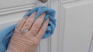 A house cleaner's gloved hand wiping a white kitchen cabinet with a blue cloth for Maid For You Cleaning & Organizing in Peoria, IL.