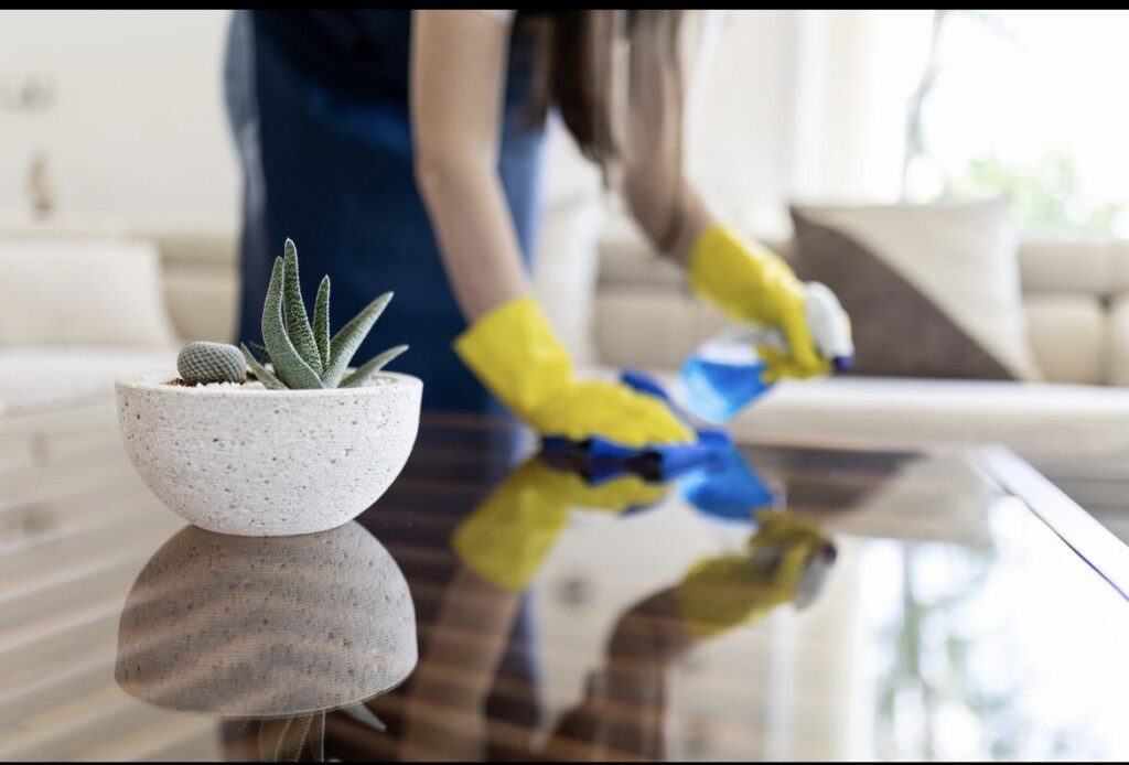 A house cleaner from MJ Commercial & Residential Cleaning, LLC wearing yellow gloves, wiping a glass coffee table in Cedar Rapids, IA.