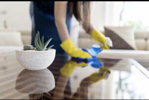 A house cleaner from MJ Commercial & Residential Cleaning, LLC wearing yellow gloves, wiping a glass coffee table in Cedar Rapids, IA.