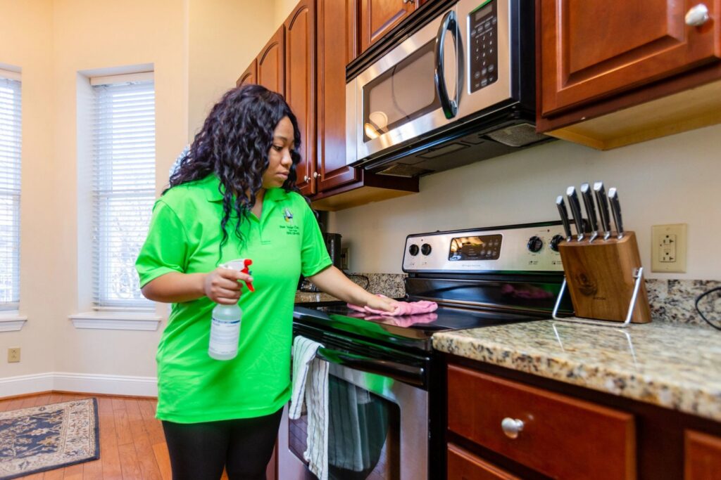 A house cleaner in a green shirt wiping down a kitchen stovetop with a cloth and spray bottle by New Image Cleaning Service LLC in Richmond, VA.
