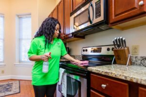 A house cleaner in a green shirt wiping down a kitchen stovetop with a cloth and spray bottle by New Image Cleaning Service LLC in Richmond, VA.