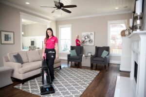 Two house cleaners from Two Maids of Hilton Head, SC, vacuuming a rug and dusting in a spacious living room.