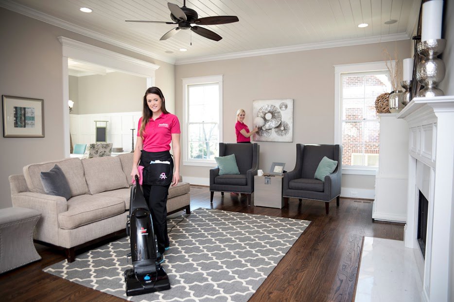 Two house cleaners from Two Maids of Hilton Head, SC, vacuuming a rug and dusting in a spacious living room.