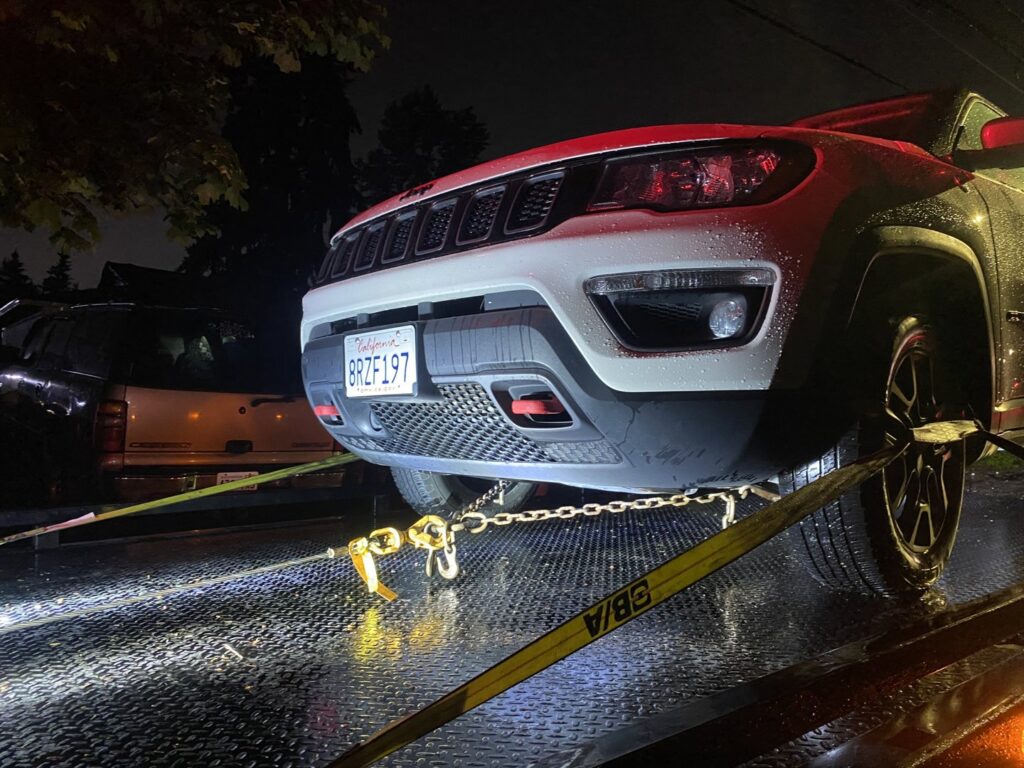 A white Jeep Compass securely loaded on a flatbed tow truck from Safety AUTO Towing LLC in Seattle, WA, at night.