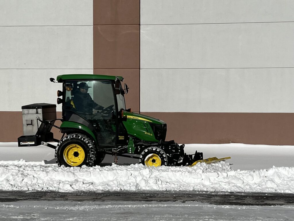 A green John Deere tractor with a snow plow attachment actively clearing snow for Appleton Snow Removal in Appleton, WI.