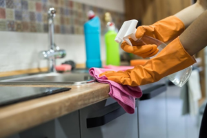 Hands in orange gloves spray and wipe a kitchen counter next to a sink for Glory Cleaning Service in Irvine, CA.