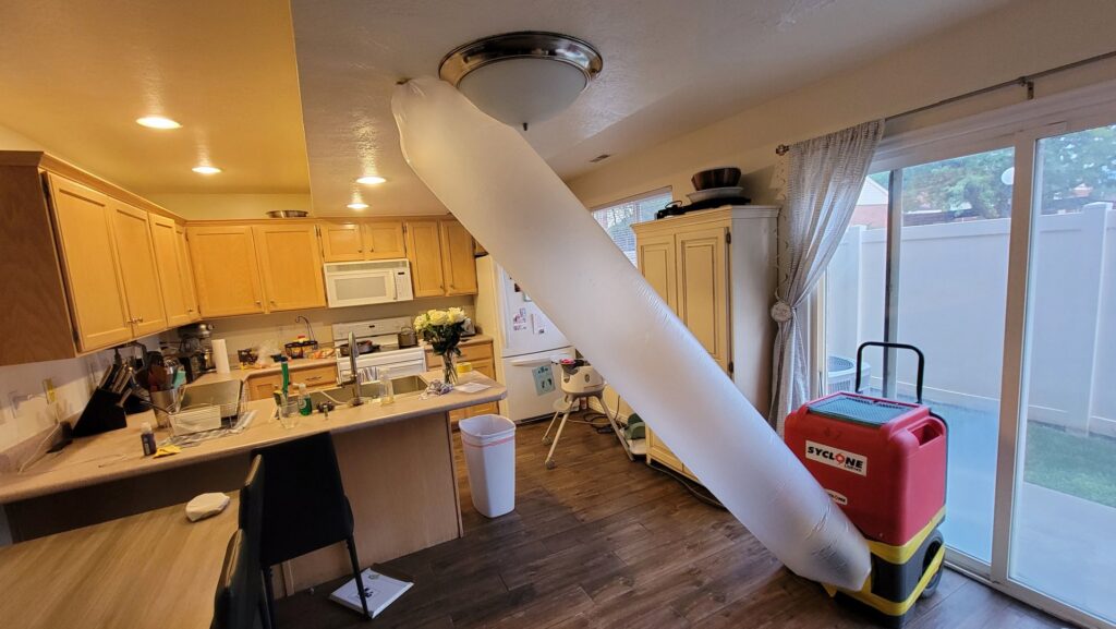 Professional drying equipment, including a large air duct and dehumidifier, set up in a kitchen for water damage restoration by Utah Disaster Team in Layton, UT.