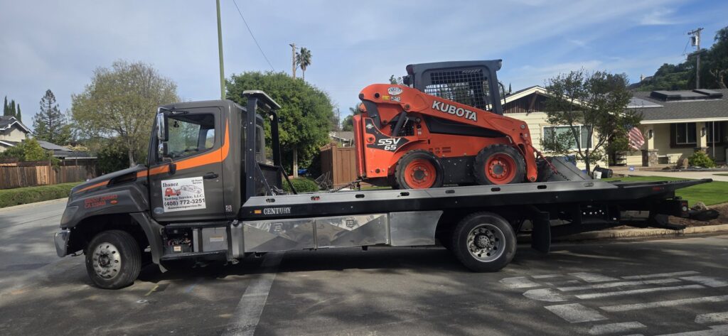 An orange Kubota skid steer loader being transported on a flatbed tow truck by Ibanez Towing Service LLC in San Jose, CA.