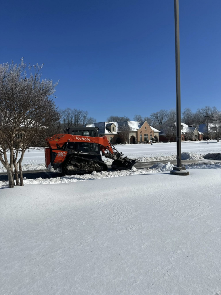 A Kubota skid steer actively plowing snow in a parking lot, demonstrating snow removal services by Hays Landscape Solutions LLC in Jackson, TN.