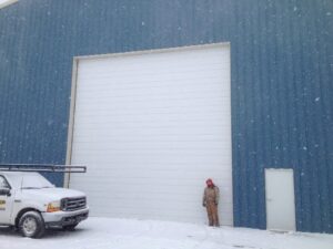 A large white commercial garage door installed on a blue metal building with a worker and service truck in the snow by Alberts Custom Door Company in Wichita, KS.