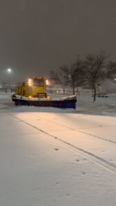 A large yellow snow plow loader parked in a snow-covered commercial parking lot at night, ready for work by Appleton Snow Removal in Appleton, WI.