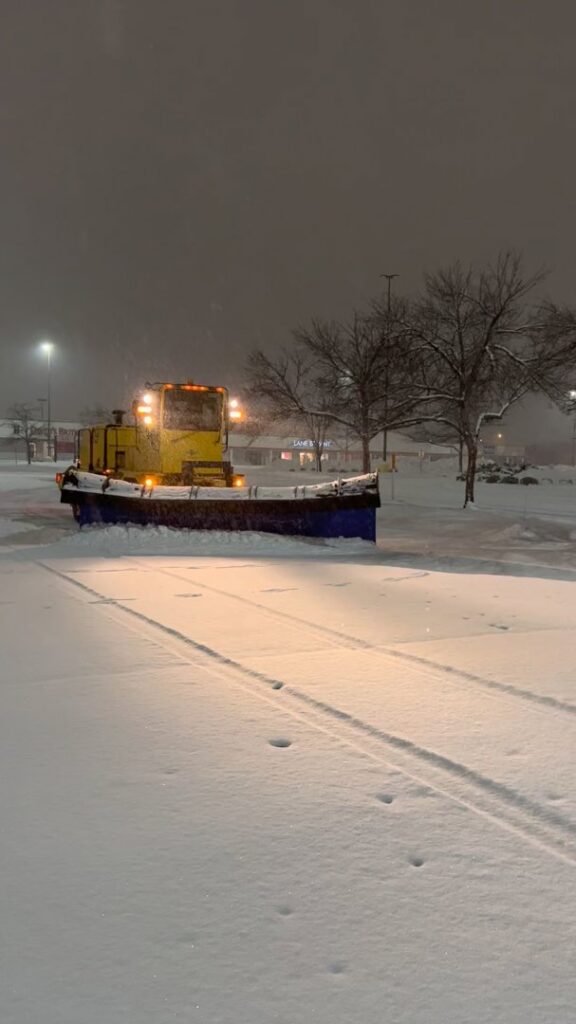 A large yellow snow plow loader parked in a snow-covered commercial parking lot at night, ready for work by Appleton Snow Removal in Appleton, WI.